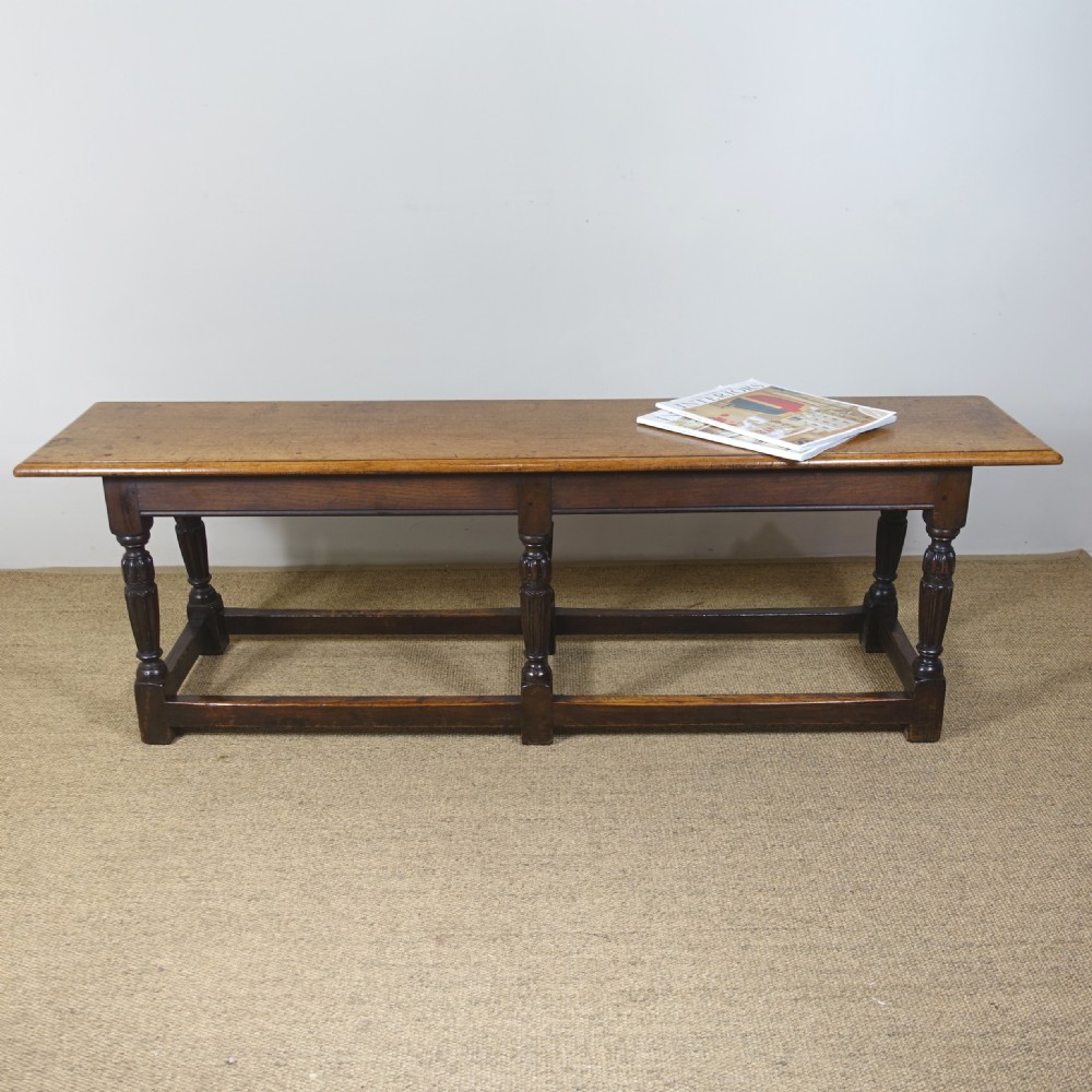 A long 19th-century dark oak stool featuring a flat plank top supported by six turned legs and low perimeter stretchers. View VII