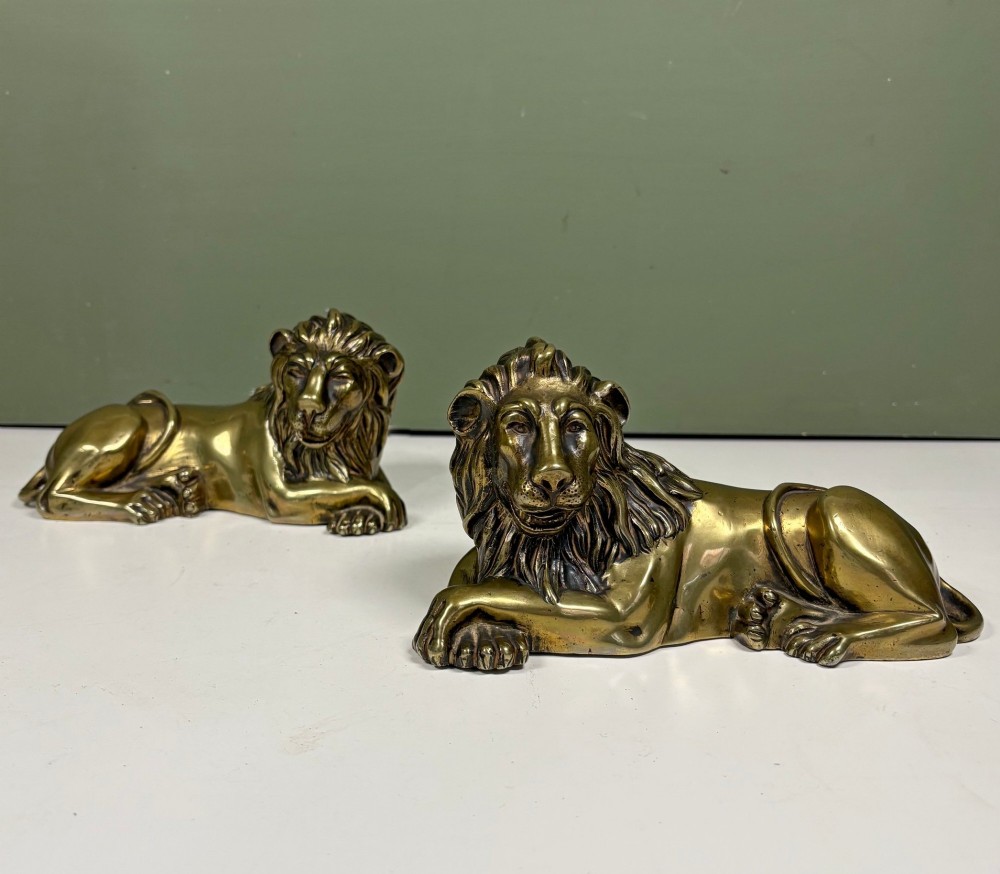 A pair of cast brass figures depicting recumbent lions with detailed manes and crossed paws facing forward.