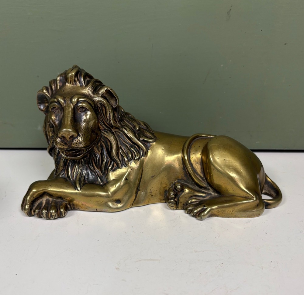 A pair of cast brass figures depicting recumbent lions with detailed manes and crossed paws facing forward. View II