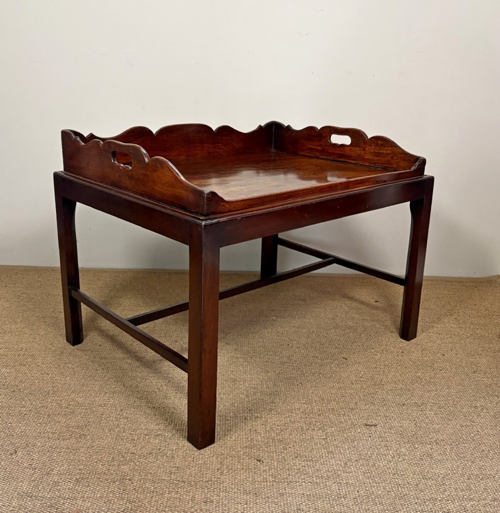 A George III mahogany butlers tray table featuring a scalloped gallery edge with cut-out handles supported by a square-leg wooden stand with cross stretchers.