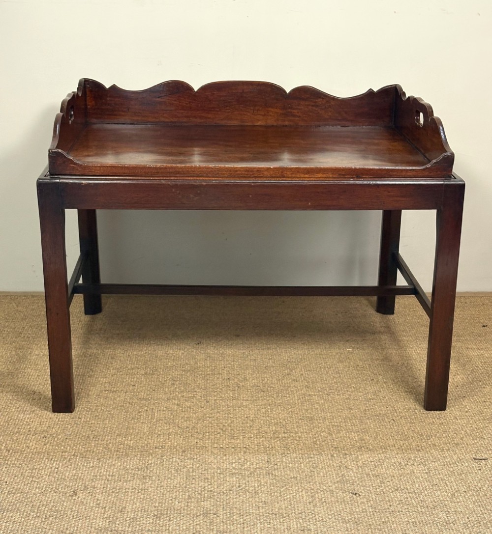 A George III mahogany butlers tray table featuring a scalloped gallery edge with cut-out handles supported by a square-leg wooden stand with cross stretchers. View II