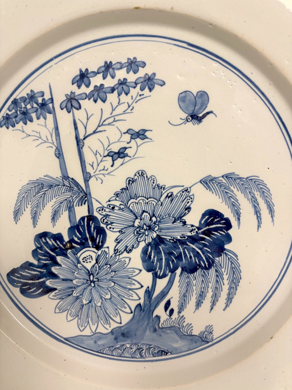 An 18th-century circular blue and white tin-glazed earthenware charger featuring a central hand-painted floral motif with a butterfly detail. View V