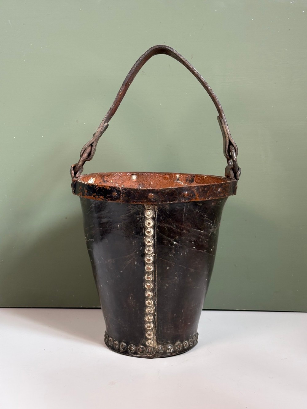 A 19th-century dark brown boiled leather bucket featuring a vertical row of copper rivets down the seam and a metal-reinforced leather strap handle.
