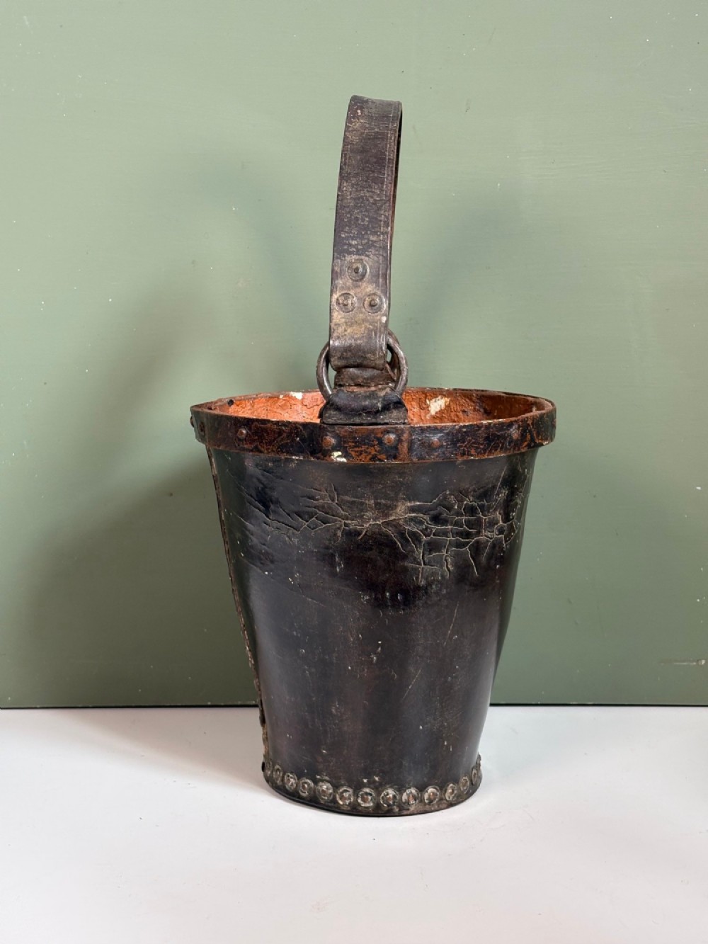 A 19th-century dark brown boiled leather bucket featuring a vertical row of copper rivets down the seam and a metal-reinforced leather strap handle. View III
