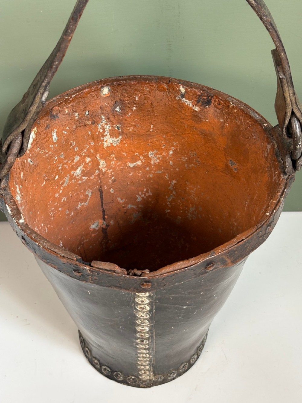 A 19th-century dark brown boiled leather bucket featuring a vertical row of copper rivets down the seam and a metal-reinforced leather strap handle. View IV