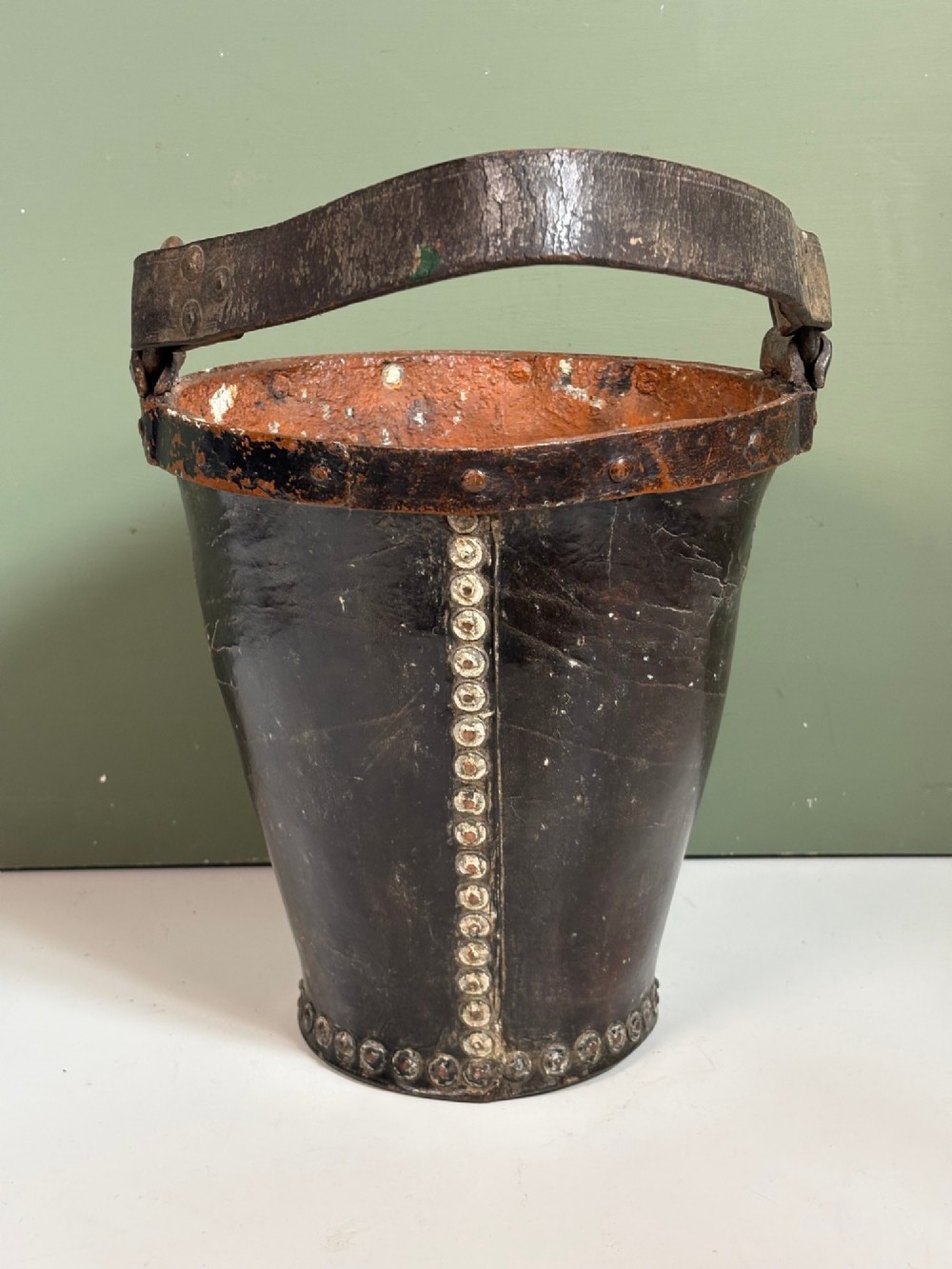 A 19th-century dark brown boiled leather bucket featuring a vertical row of copper rivets down the seam and a metal-reinforced leather strap handle. View V