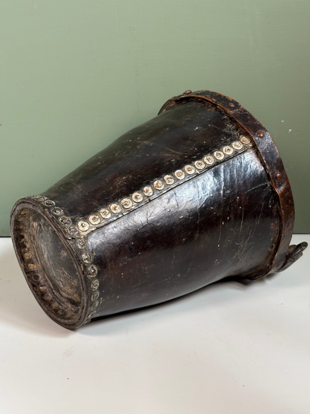 A 19th-century dark brown boiled leather bucket featuring a vertical row of copper rivets down the seam and a metal-reinforced leather strap handle. View VI