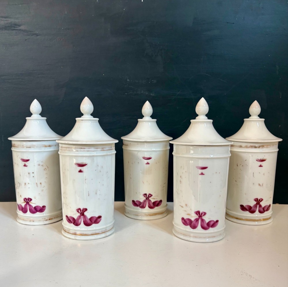 Set of five matching off-white 19th-century porcelain apothecary jars featuring pointed finial lids and hand-painted magenta bow motifs.
