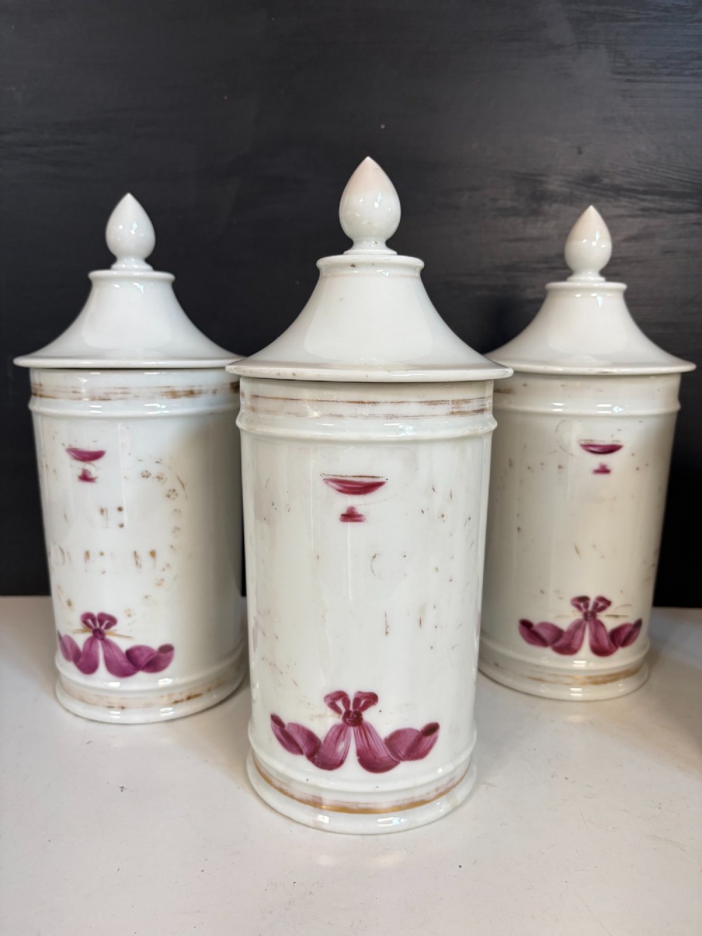 Set of five matching off-white 19th-century porcelain apothecary jars featuring pointed finial lids and hand-painted magenta bow motifs. View II
