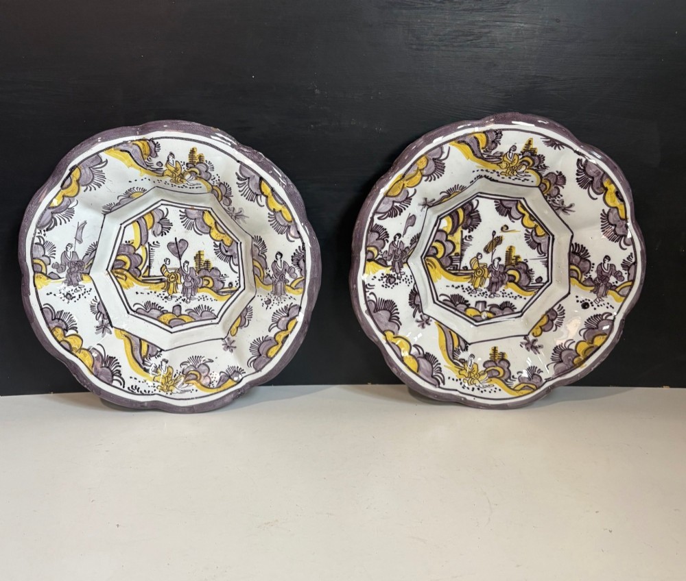A pair of circular 18th-century tin-glazed earthenware dishes featuring hand-painted manganese purple and yellow chinoiserie figures surrounded by scalloped edges.