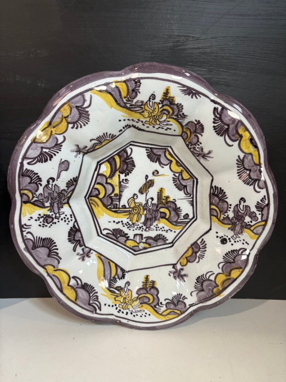 A pair of circular 18th-century tin-glazed earthenware dishes featuring hand-painted manganese purple and yellow chinoiserie figures surrounded by scalloped edges. View II