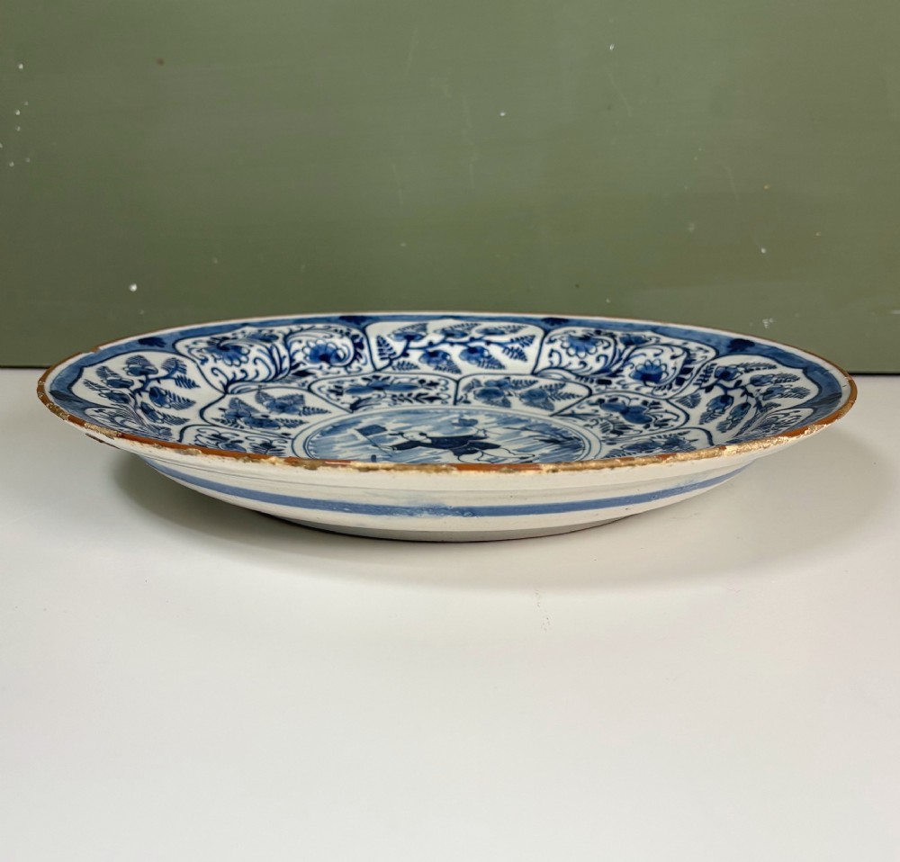 A circular 18th-century tin-glazed earthenware dish decorated in cobalt blue and white with a central Chinoiserie figure surrounded by floral border panels. View V