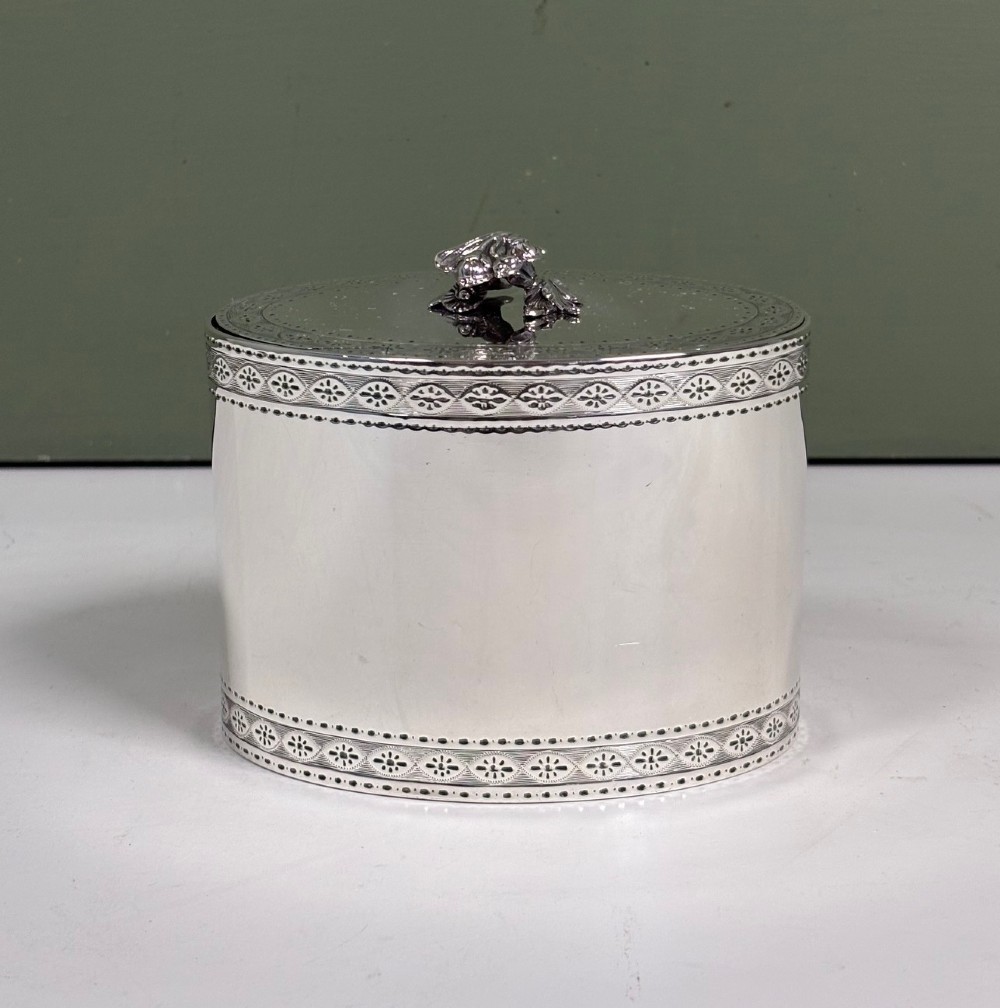 An oval sterling silver tea caddy featuring bright-cut engraved borders, visible hallmarks, and a lid crowned with a sculpted bird finial. View II