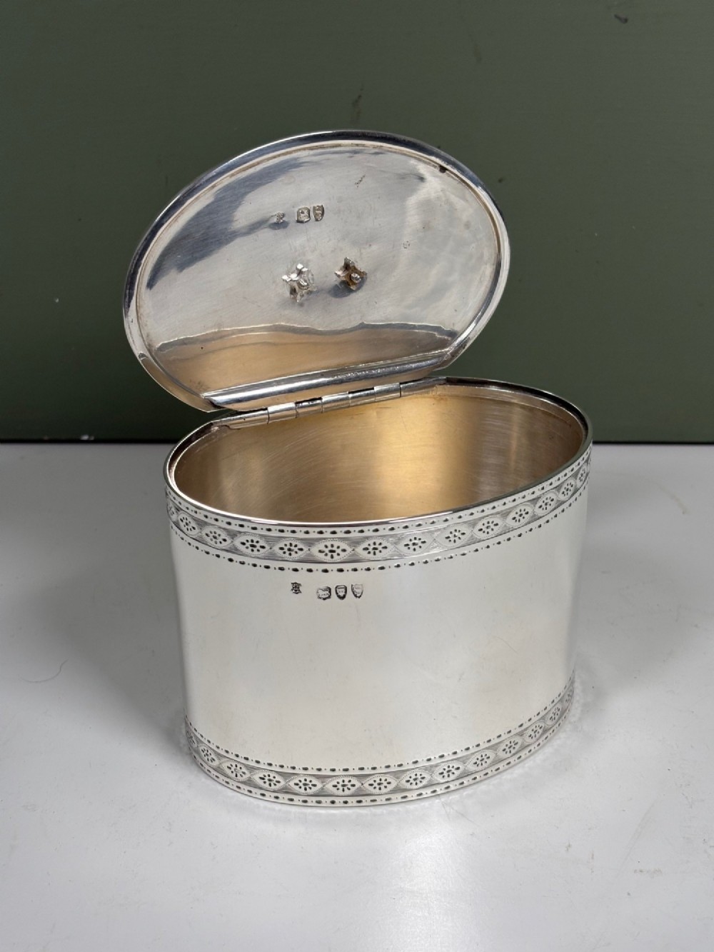 An oval sterling silver tea caddy featuring bright-cut engraved borders, visible hallmarks, and a lid crowned with a sculpted bird finial. View V