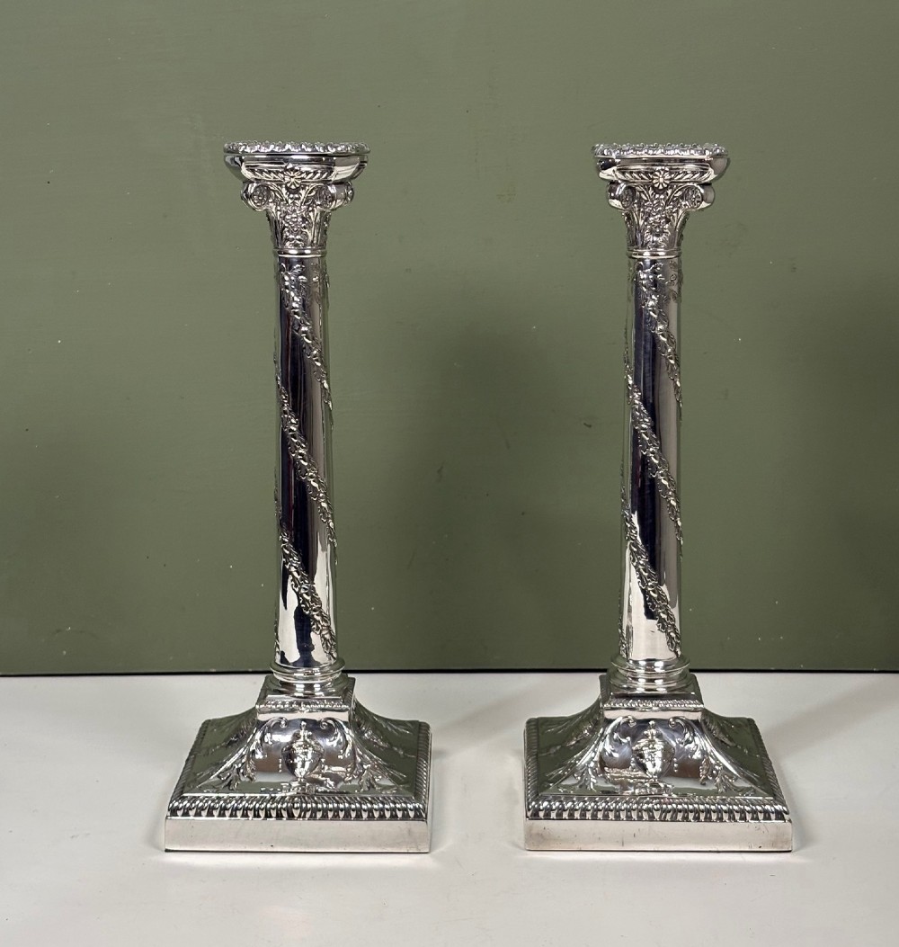 A pair of 19th-century silver-plated candlesticks featuring Corinthian capitals, spiral garland columns, and square stepped bases with classical urn reliefs.