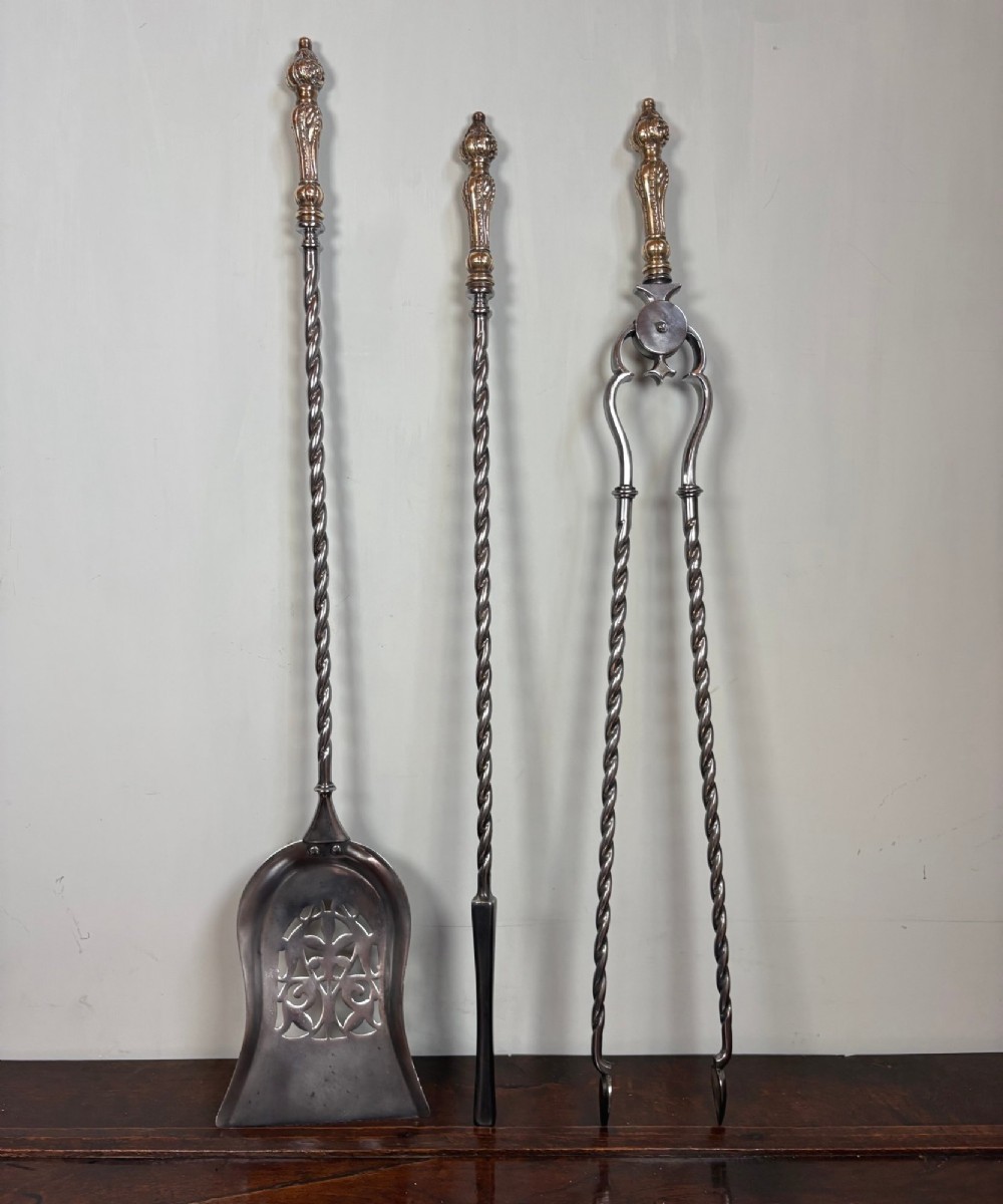A set of three 19th-century polished steel fire tools including a pierced shovel, poker, and tongs with barley-twist shafts and ornate brass handles.