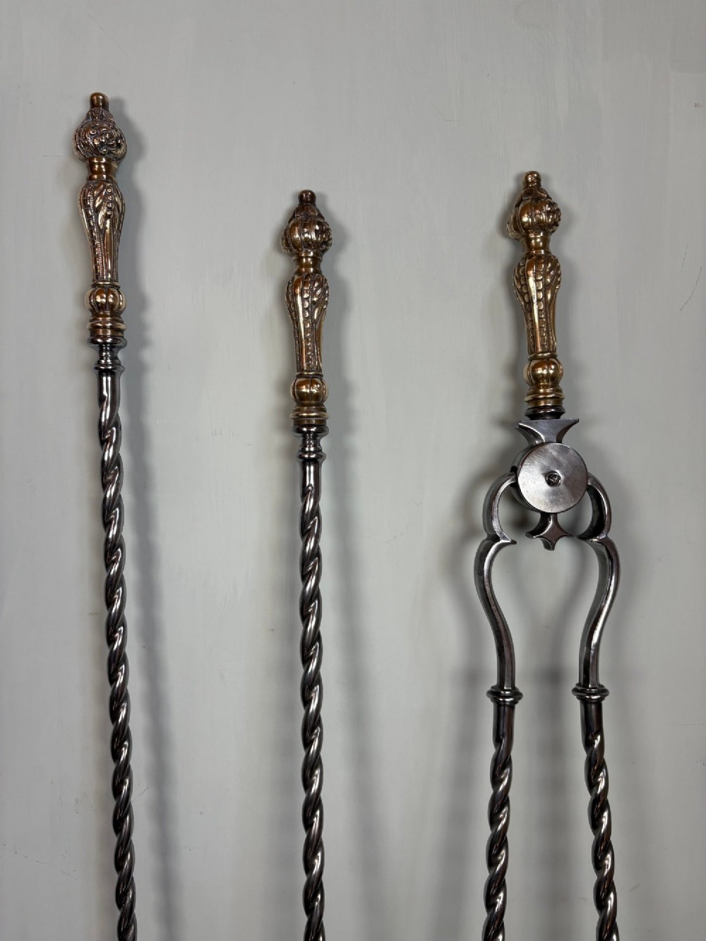 A set of three 19th-century polished steel fire tools including a pierced shovel, poker, and tongs with barley-twist shafts and ornate brass handles. View II