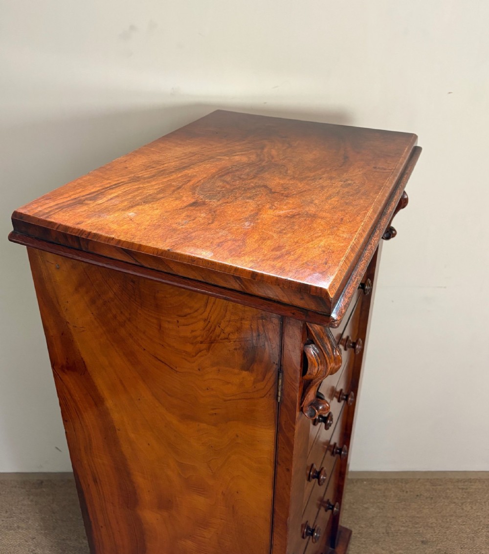 A tall Victorian walnut Wellington chest featuring six drawer fronts, carved corbels, and an open drop-front secretaire revealing internal pigeonholes and small drawers. View VI