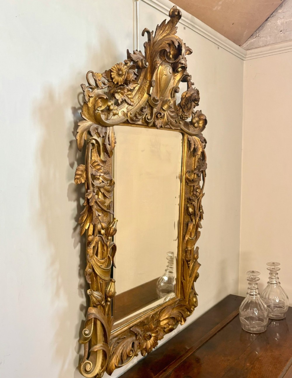 A 19th-century Rococo Revival carved giltwood wall mirror featuring ornate floral cresting, acanthus leaf details, and a central mirror plate. View VI