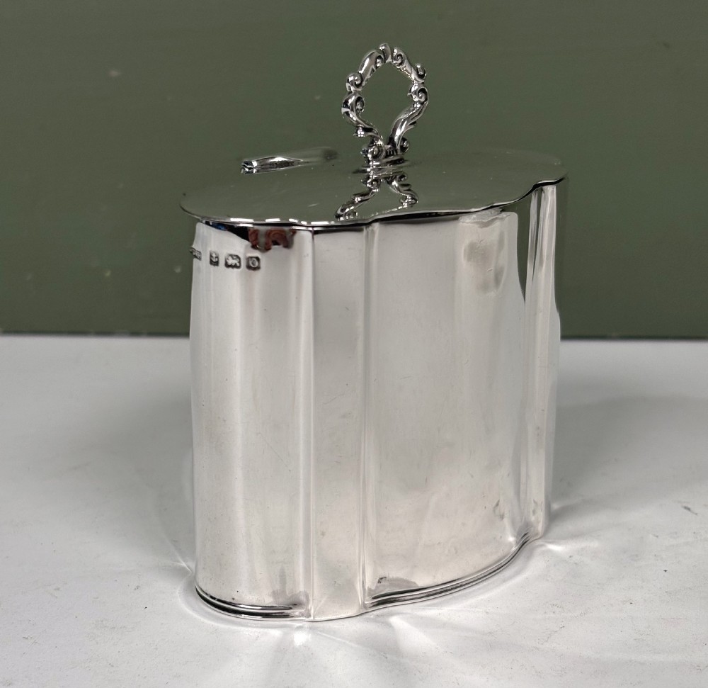A polished cylindrical solid silver caddy featuring lobed vertical panels, a fitted flush lid, and an ornate scroll finial handle. View II