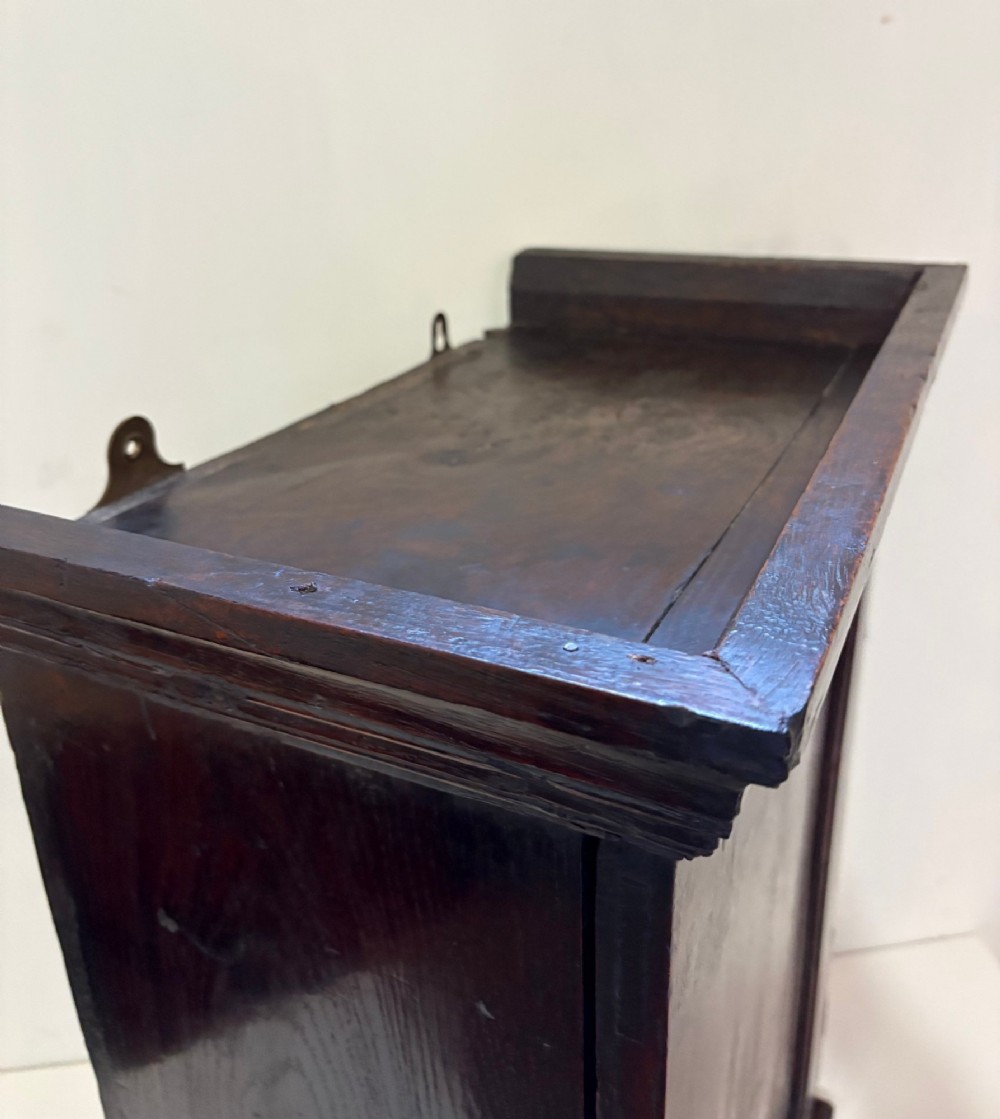 An eighteenth-century dark oak wall cupboard with a single field-panel door, brass keyhole escutcheon, and moulded top cornice. View V