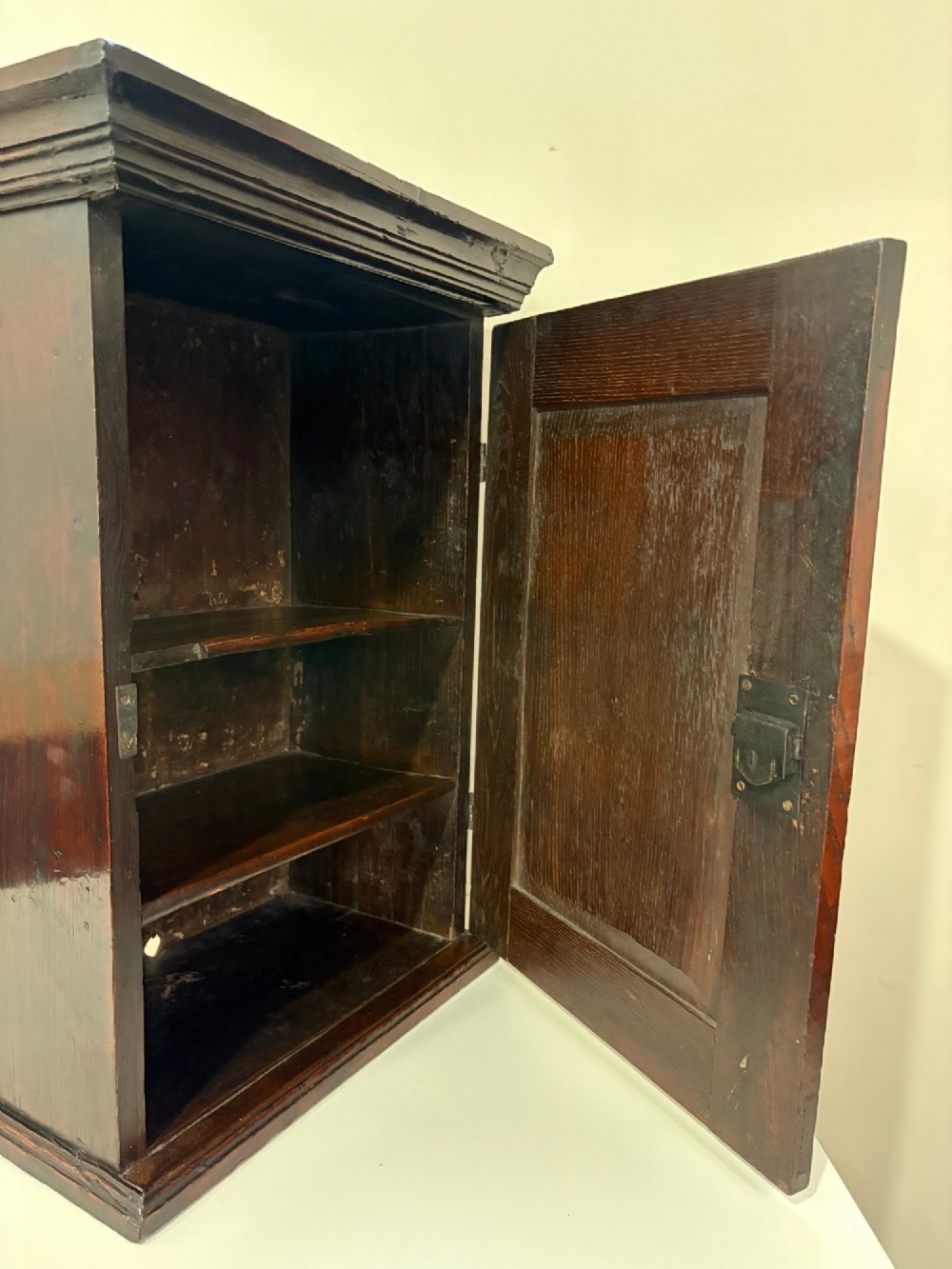 An eighteenth-century dark oak wall cupboard with a single field-panel door, brass keyhole escutcheon, and moulded top cornice. View VII