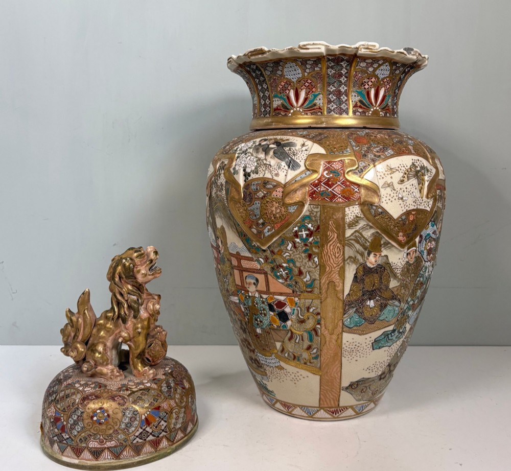 A large Japanese Satsuma earthenware lidded vase featuring intricate gilding, figural panels, a scalloped collar, and a gilded foo dog finial. View V