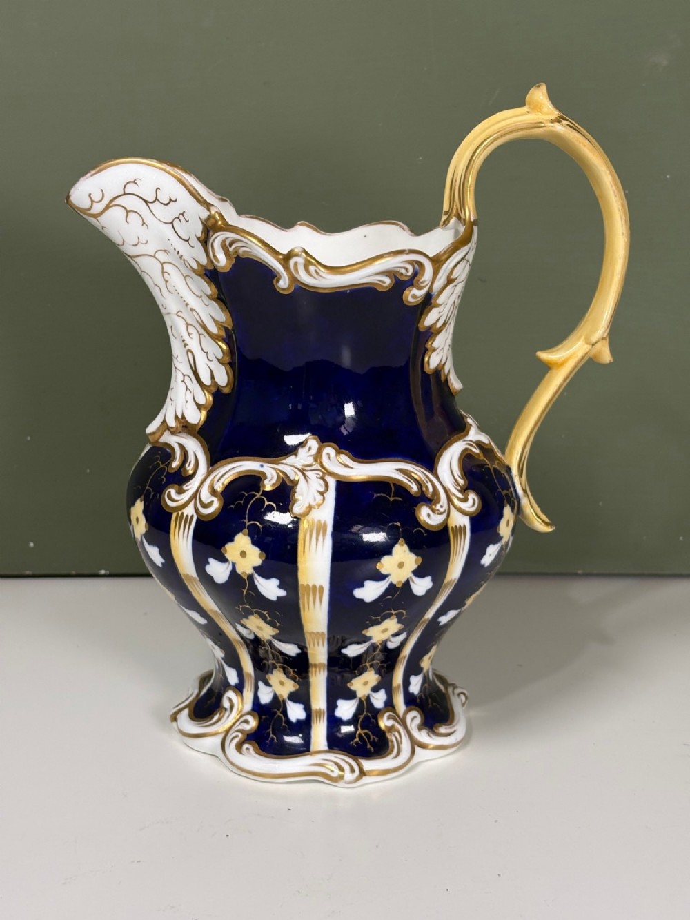 A nineteenth-century cobalt blue porcelain jug with gold gilding, white floral pattern, an ornate scroll handle, and a scalloped spout. View II
