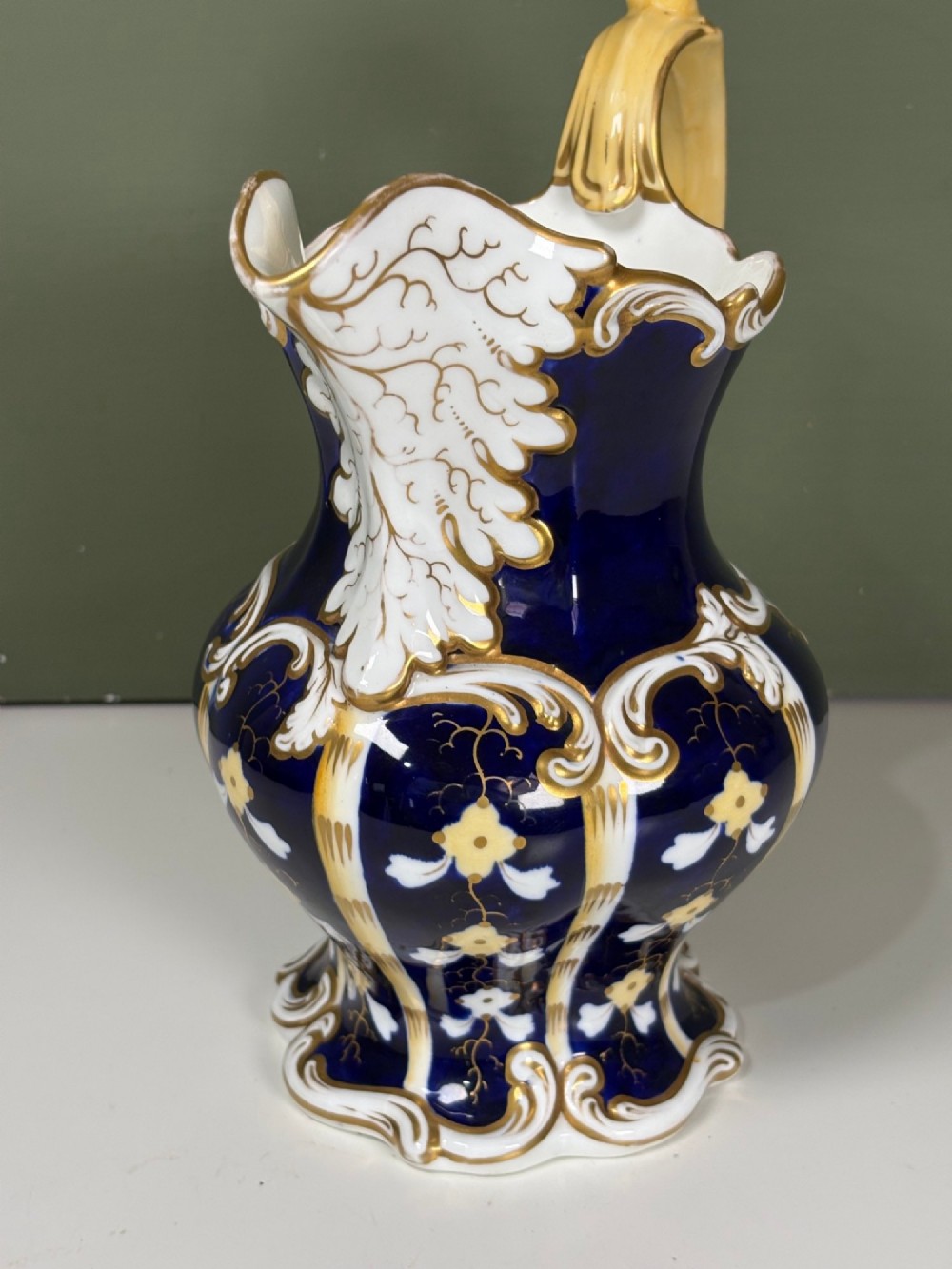 A nineteenth-century cobalt blue porcelain jug with gold gilding, white floral pattern, an ornate scroll handle, and a scalloped spout. View V
