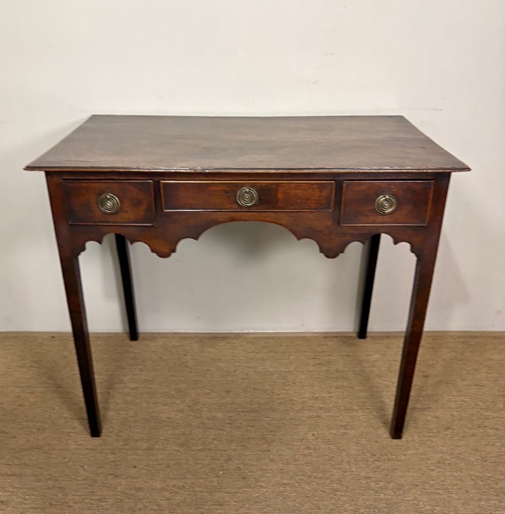 An 18th-century dark oak lowboy side table featuring three drawers with circular brass drop handles and a shaped scalloped apron supported by four straight legs. View II