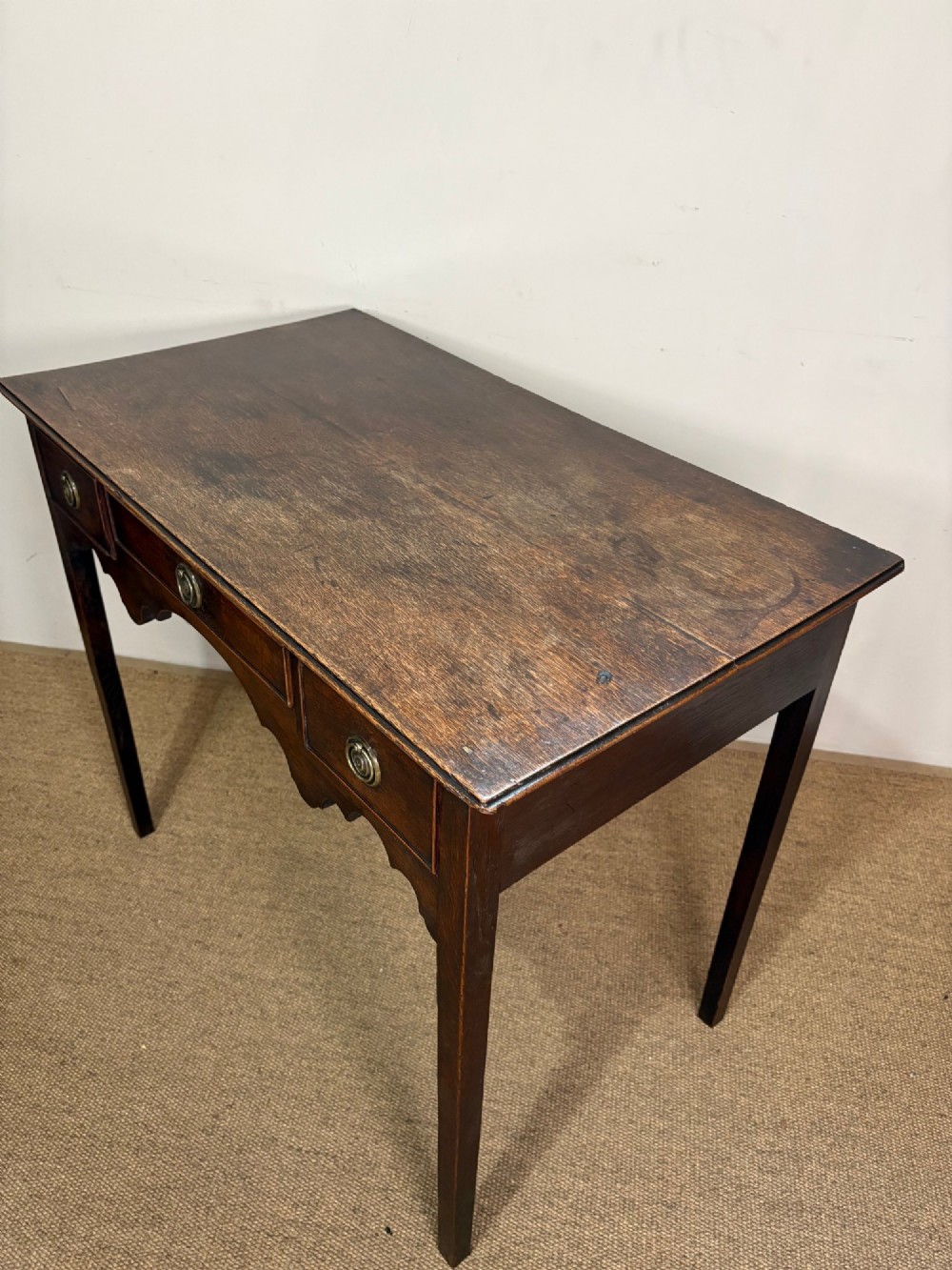 An 18th-century dark oak lowboy side table featuring three drawers with circular brass drop handles and a shaped scalloped apron supported by four straight legs. View V