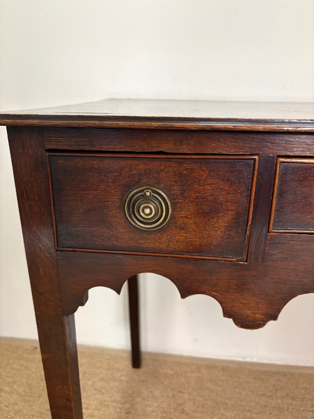 An 18th-century dark oak lowboy side table featuring three drawers with circular brass drop handles and a shaped scalloped apron supported by four straight legs. View VII