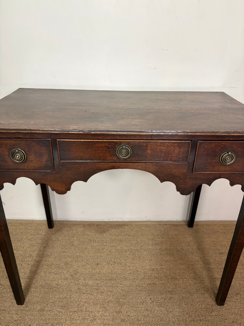 An 18th-century dark oak lowboy side table featuring three drawers with circular brass drop handles and a shaped scalloped apron supported by four straight legs. View VIII