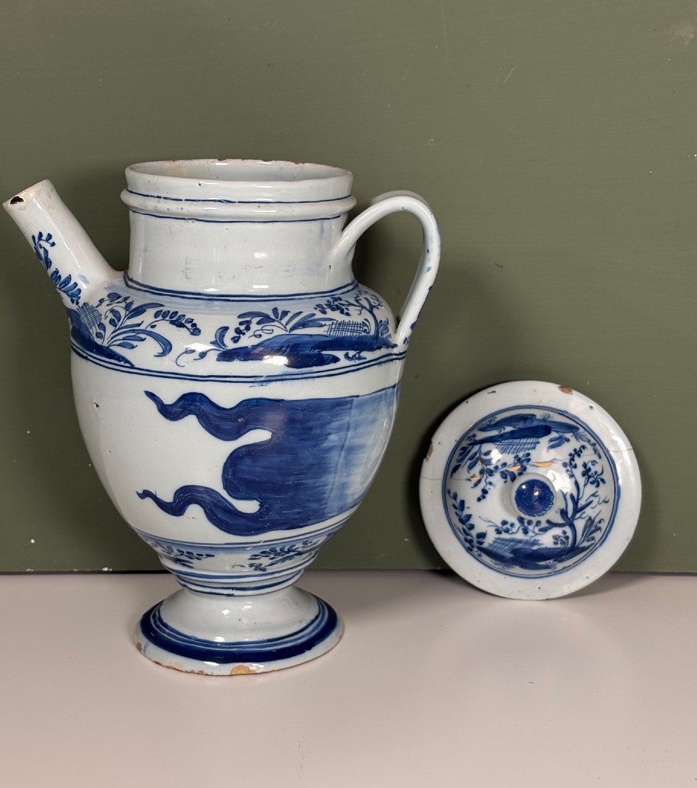 An 18th-century blue and white tin-glazed Delftware spouted apothecary jar featuring a fitted lid, side handle, and painted foliate decoration. View VI