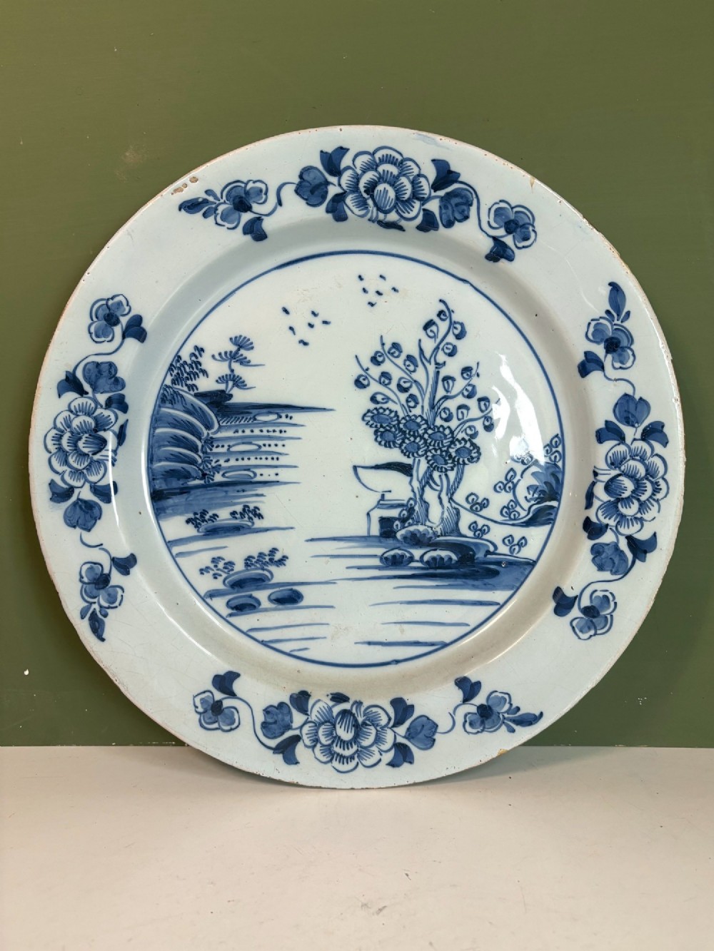 An 18th-century Dutch Delft tin-glazed earthenware charger featuring hand-painted cobalt blue floral motifs on the rim and a central chinoiserie landscape scene.