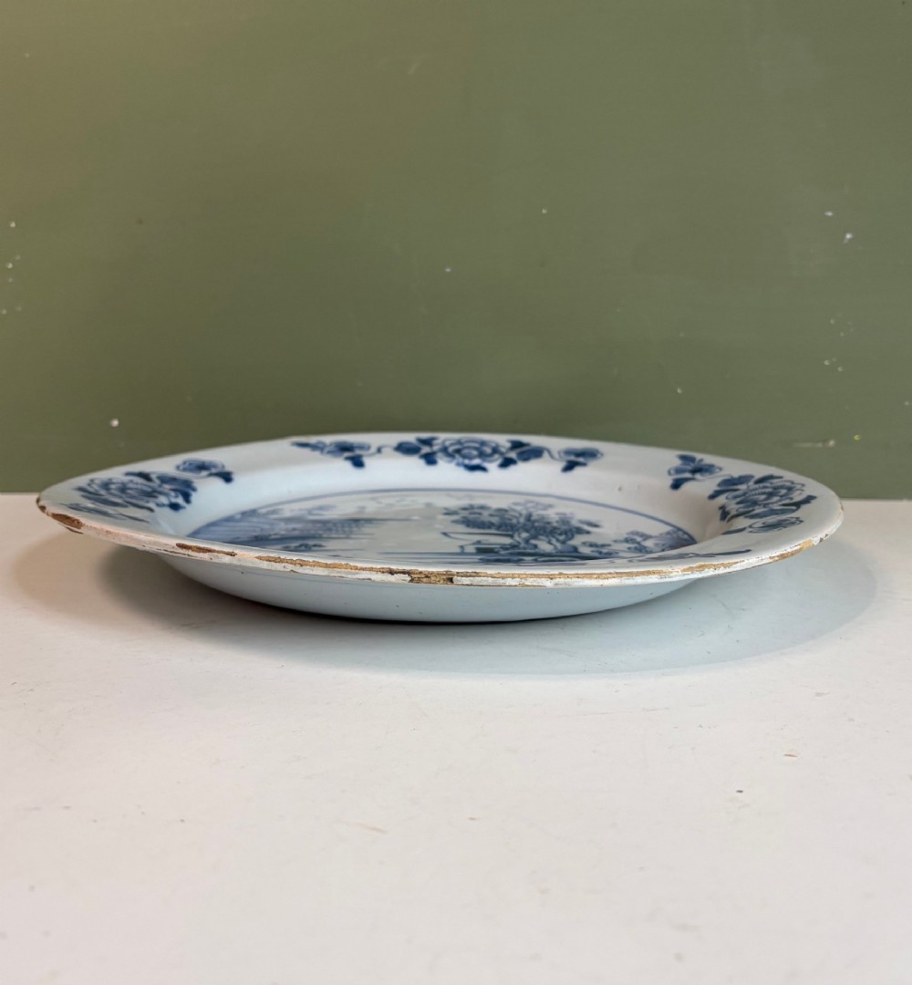 An 18th-century Dutch Delft tin-glazed earthenware charger featuring hand-painted cobalt blue floral motifs on the rim and a central chinoiserie landscape scene. View V