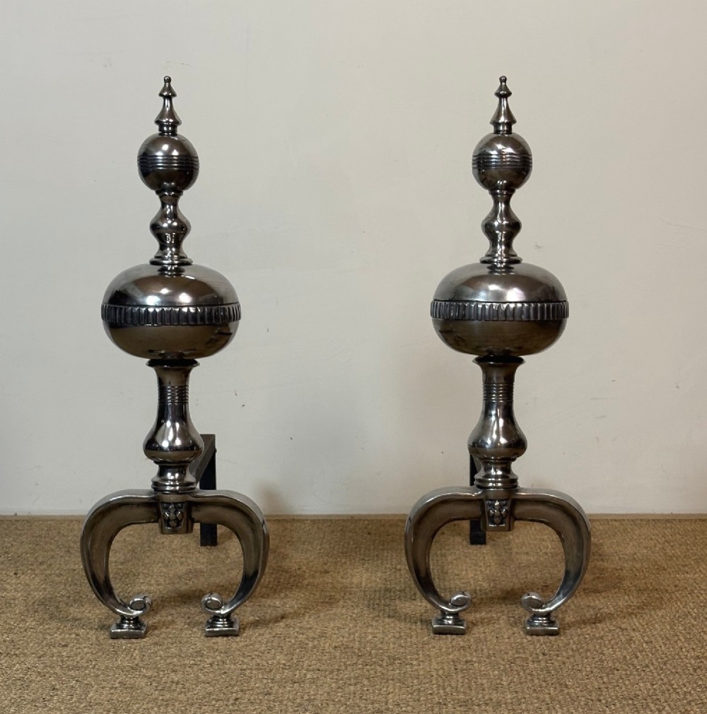 A pair of polished steel fire dogs featuring urn-shaped finials, turned stems, and arched scrolled legs.