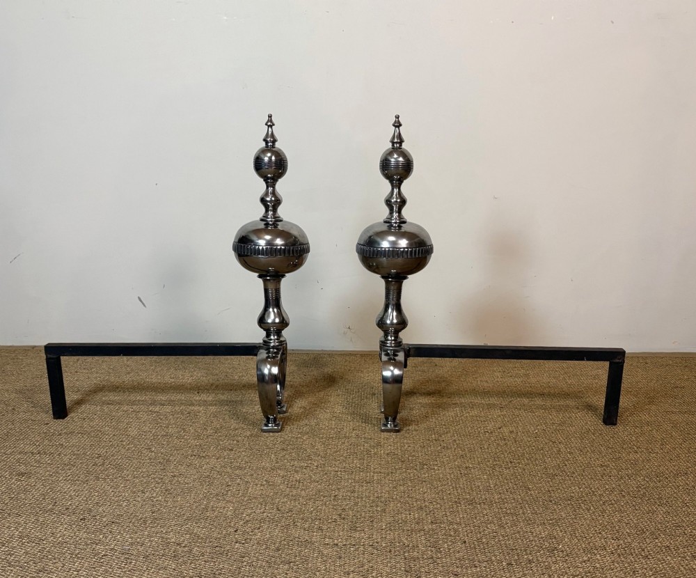 A pair of polished steel fire dogs featuring urn-shaped finials, turned stems, and arched scrolled legs. View II