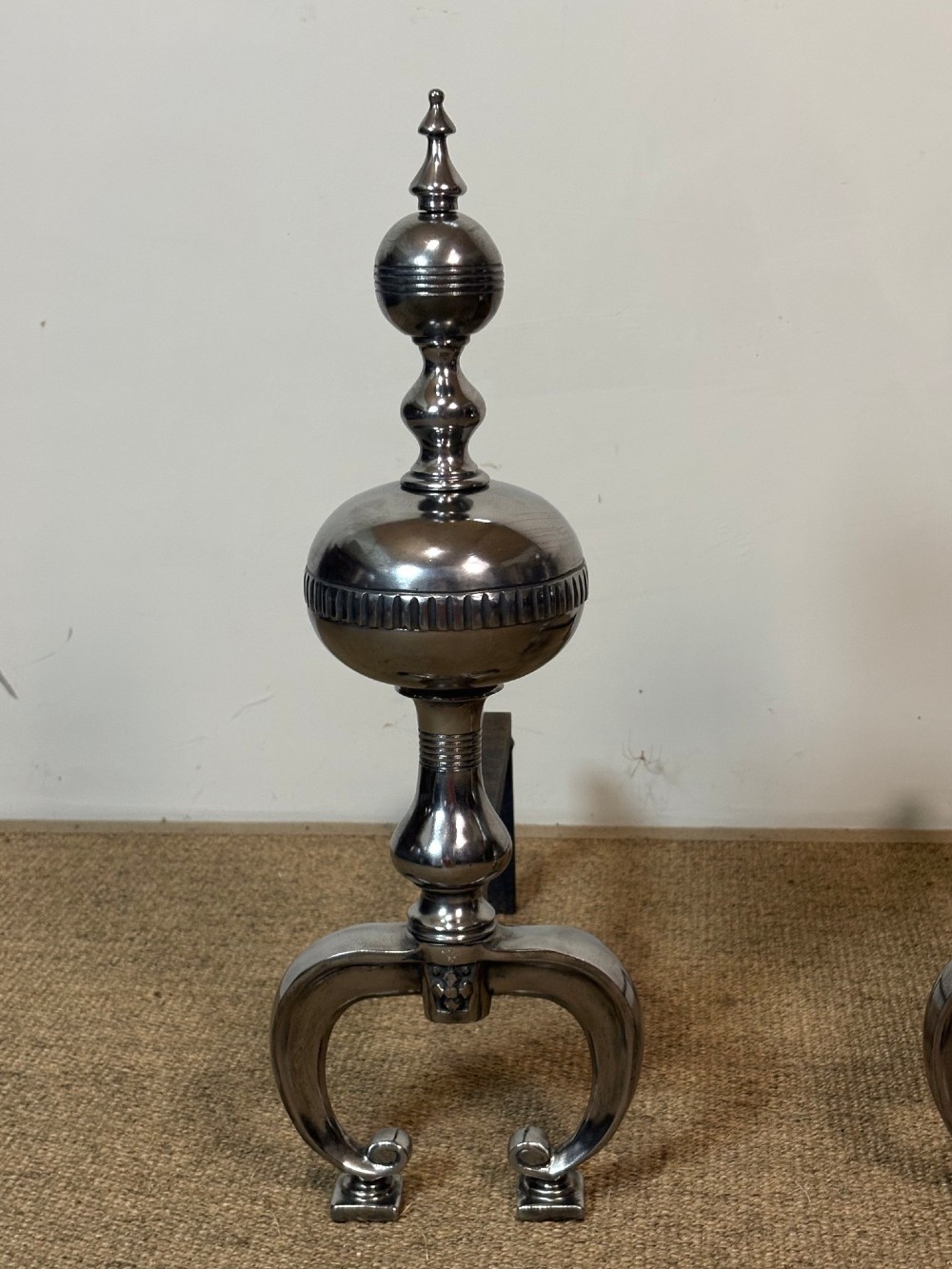 A pair of polished steel fire dogs featuring urn-shaped finials, turned stems, and arched scrolled legs. View III
