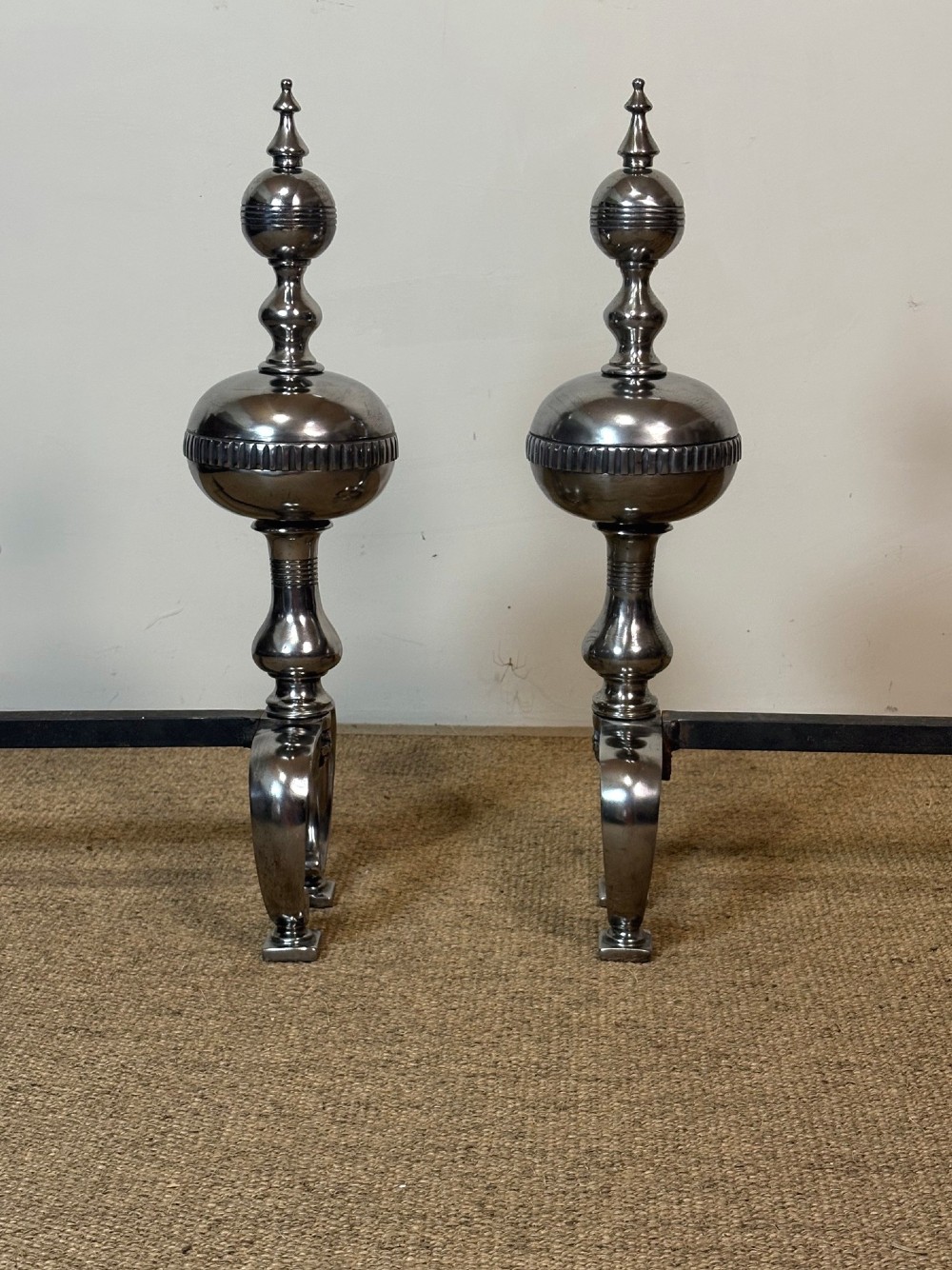 A pair of polished steel fire dogs featuring urn-shaped finials, turned stems, and arched scrolled legs. View IV