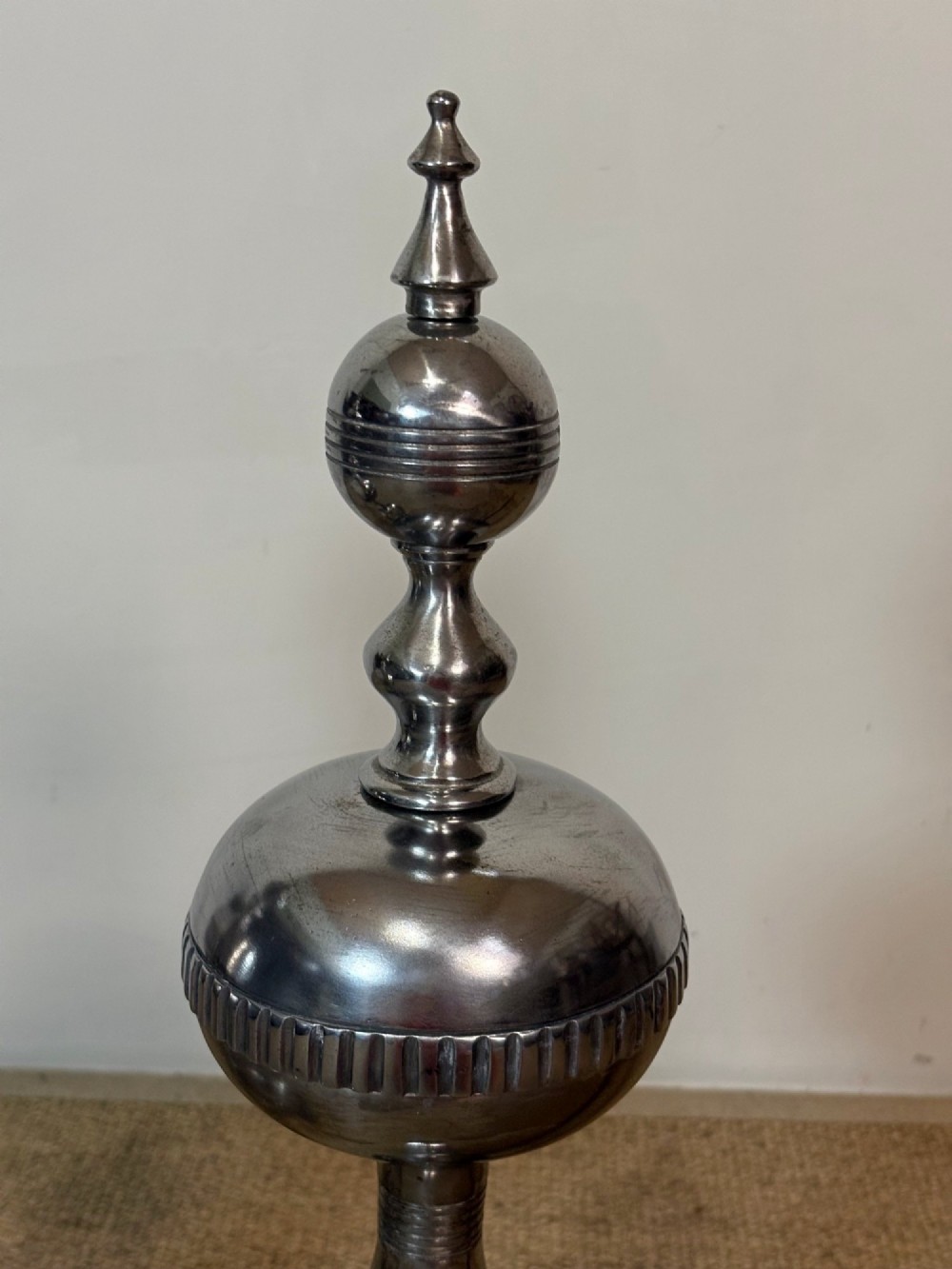 A pair of polished steel fire dogs featuring urn-shaped finials, turned stems, and arched scrolled legs. View V