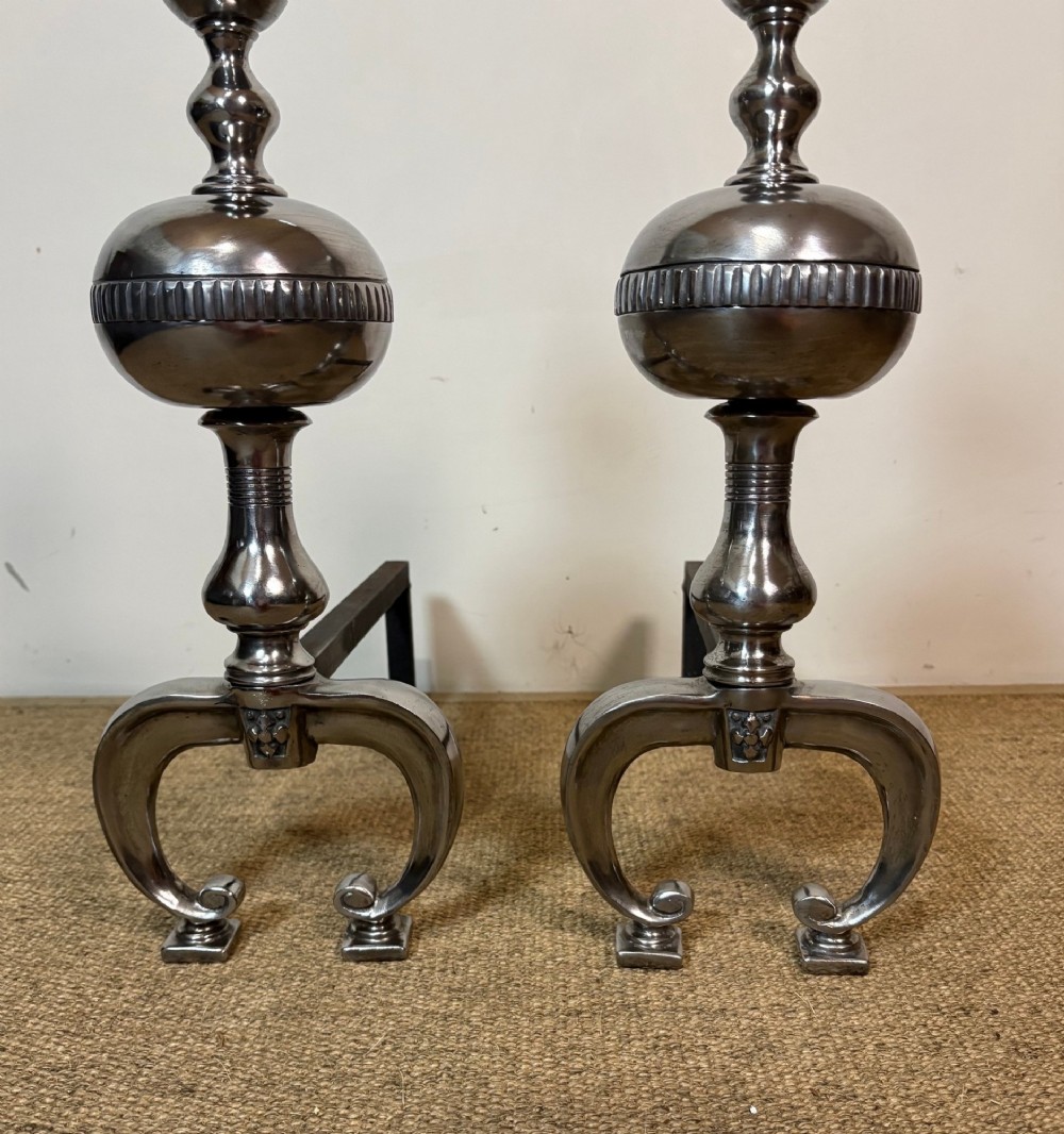 A pair of polished steel fire dogs featuring urn-shaped finials, turned stems, and arched scrolled legs. View VI