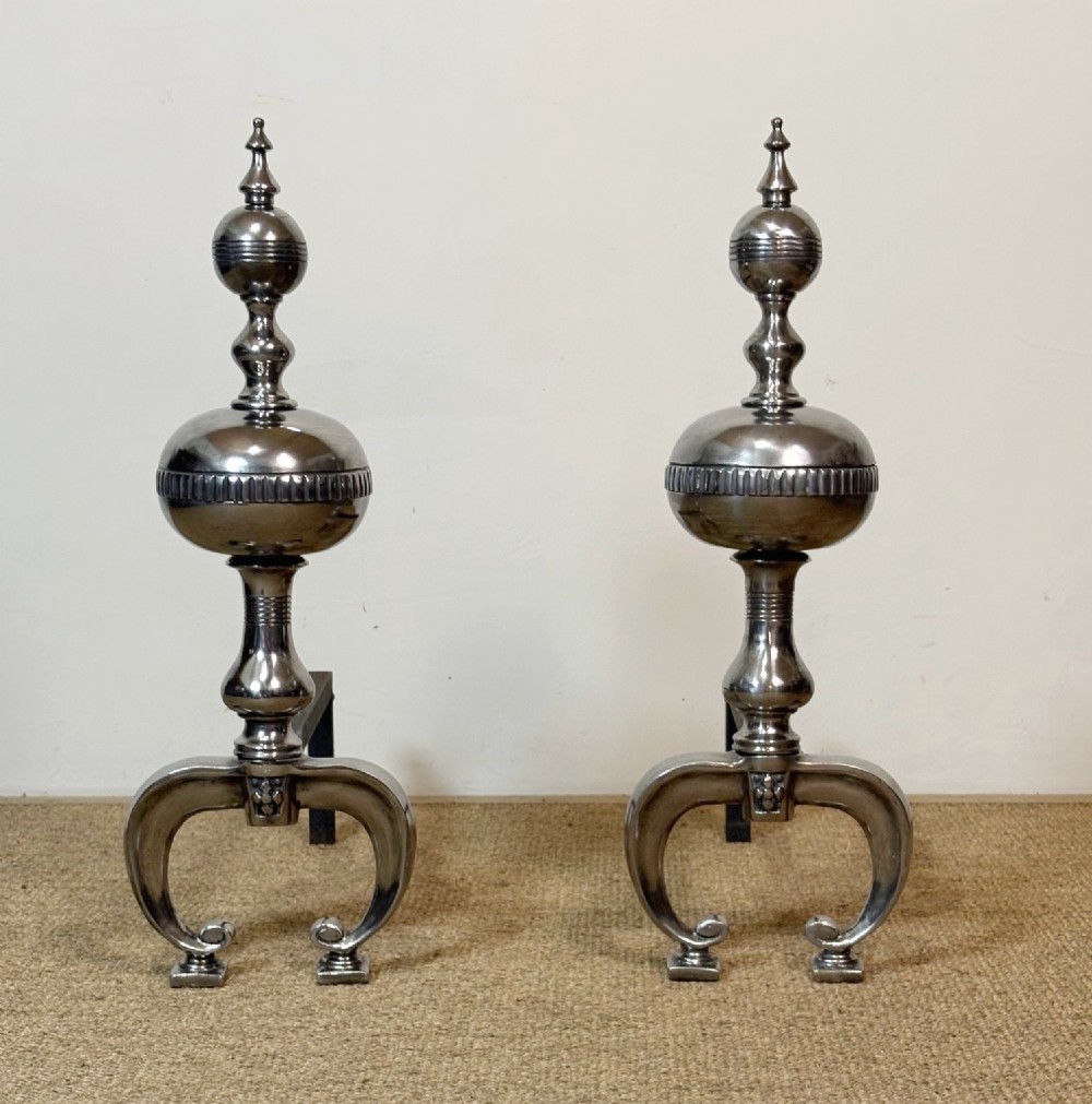 A pair of polished steel fire dogs featuring lathe-turned baluster columns, spherical fluted knops, topped with finials and supported on scrolled feet.