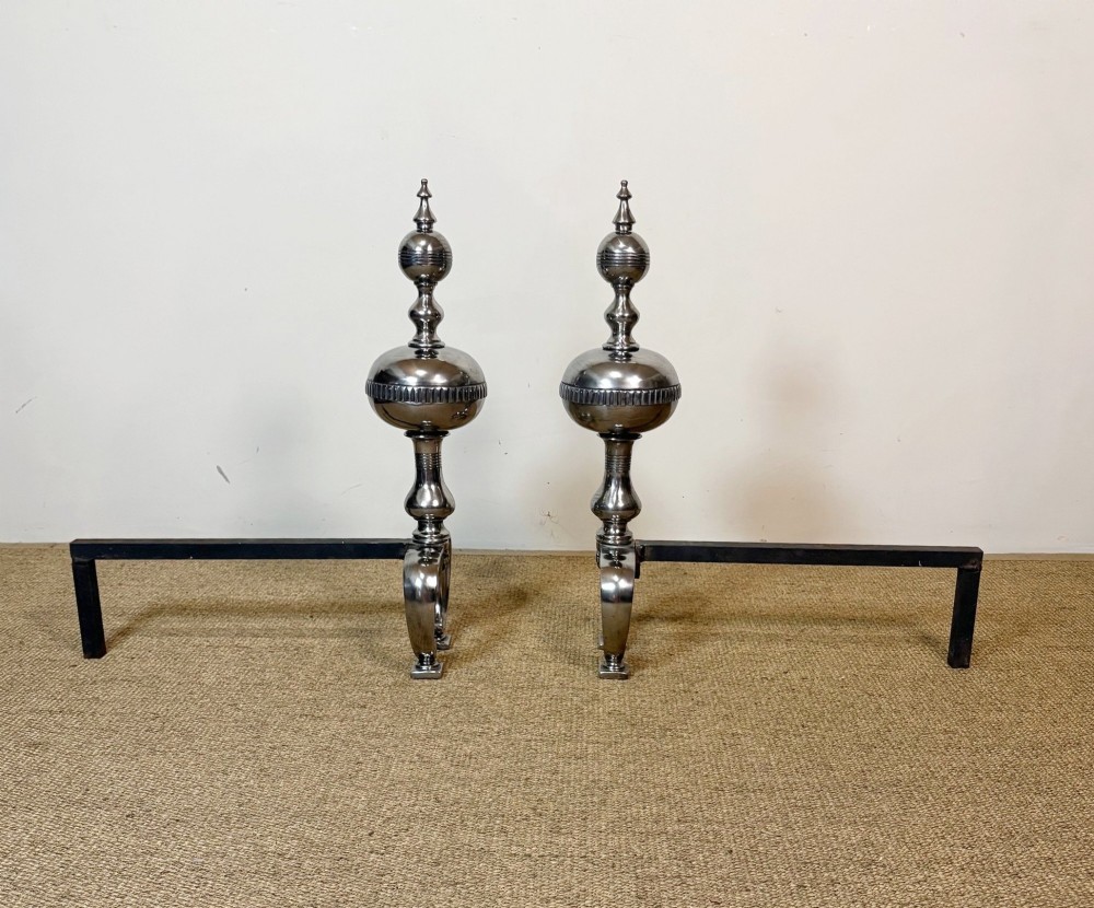 A pair of polished steel fire dogs featuring lathe-turned baluster columns, spherical fluted knops, topped with finials and supported on scrolled feet. View II