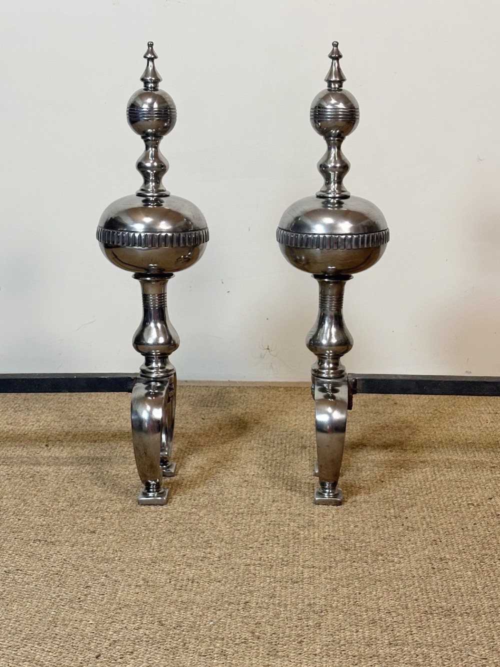 A pair of polished steel fire dogs featuring lathe-turned baluster columns, spherical fluted knops, topped with finials and supported on scrolled feet. View IV