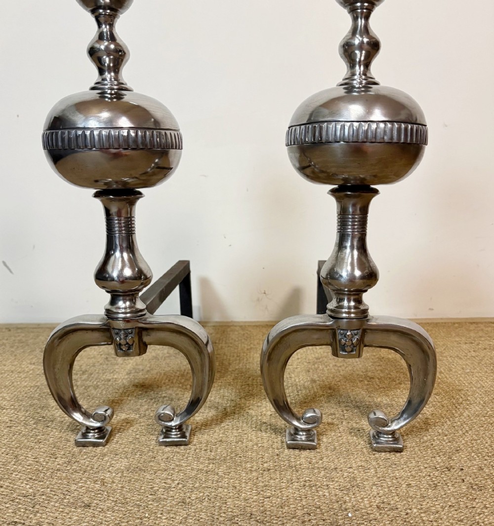 A pair of polished steel fire dogs featuring lathe-turned baluster columns, spherical fluted knops, topped with finials and supported on scrolled feet. View VI