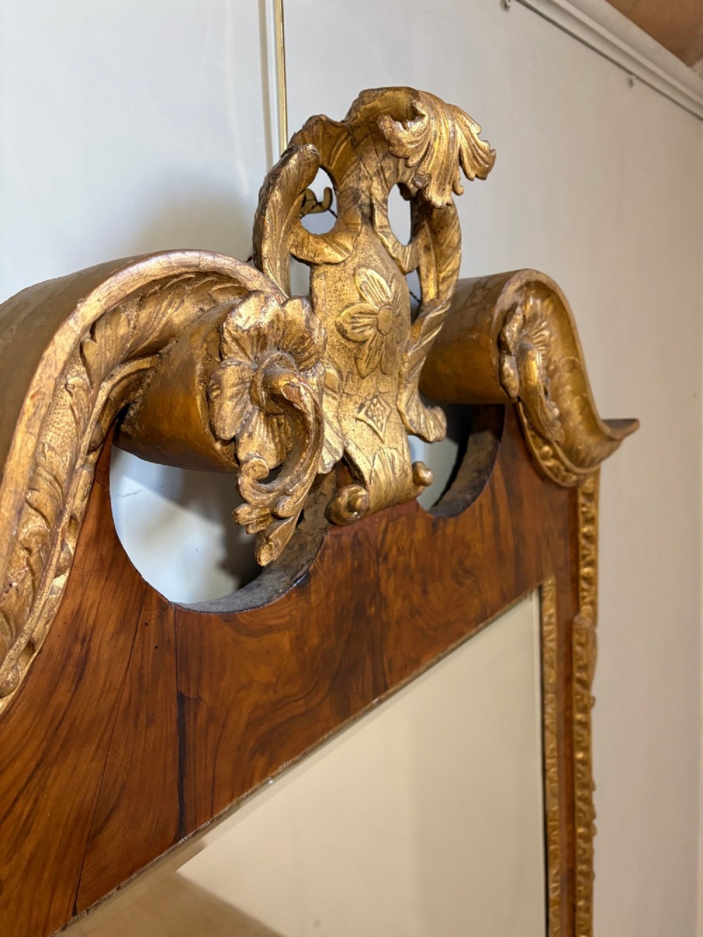 An eighteenth-century style wall mirror featuring a figured walnut veneer frame, carved giltwood foliate borders, a top cartouche cresting, and bevelled glass. View II