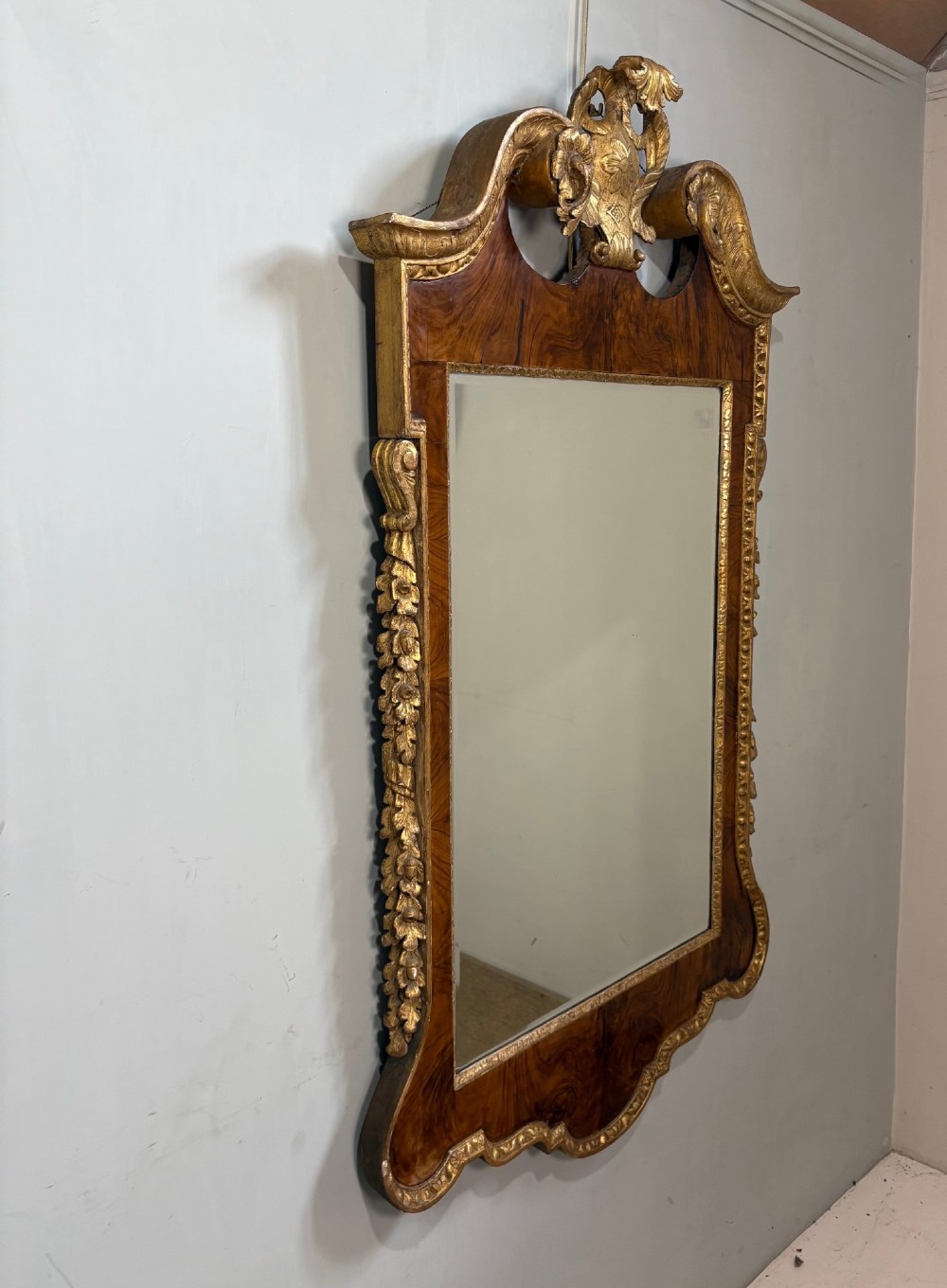 An eighteenth-century style wall mirror featuring a figured walnut veneer frame, carved giltwood foliate borders, a top cartouche cresting, and bevelled glass. View III