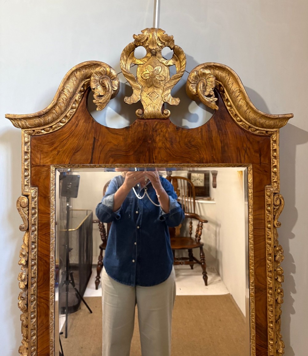 An eighteenth-century style wall mirror featuring a figured walnut veneer frame, carved giltwood foliate borders, a top cartouche cresting, and bevelled glass. View V