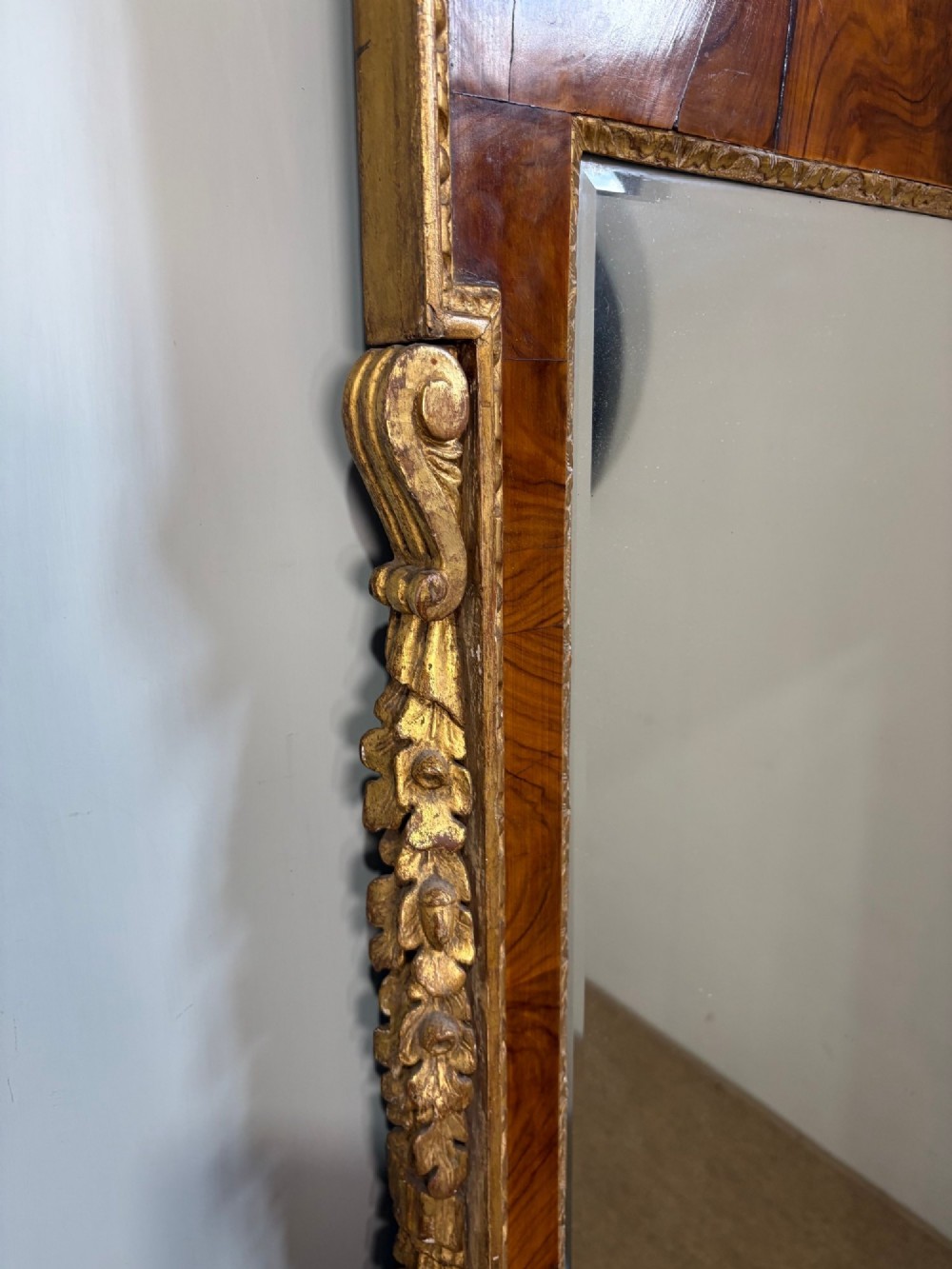An eighteenth-century style wall mirror featuring a figured walnut veneer frame, carved giltwood foliate borders, a top cartouche cresting, and bevelled glass. View VI